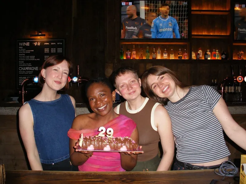 Four friends at a pub holding a caterpillar birthday cake with a 29 candle
